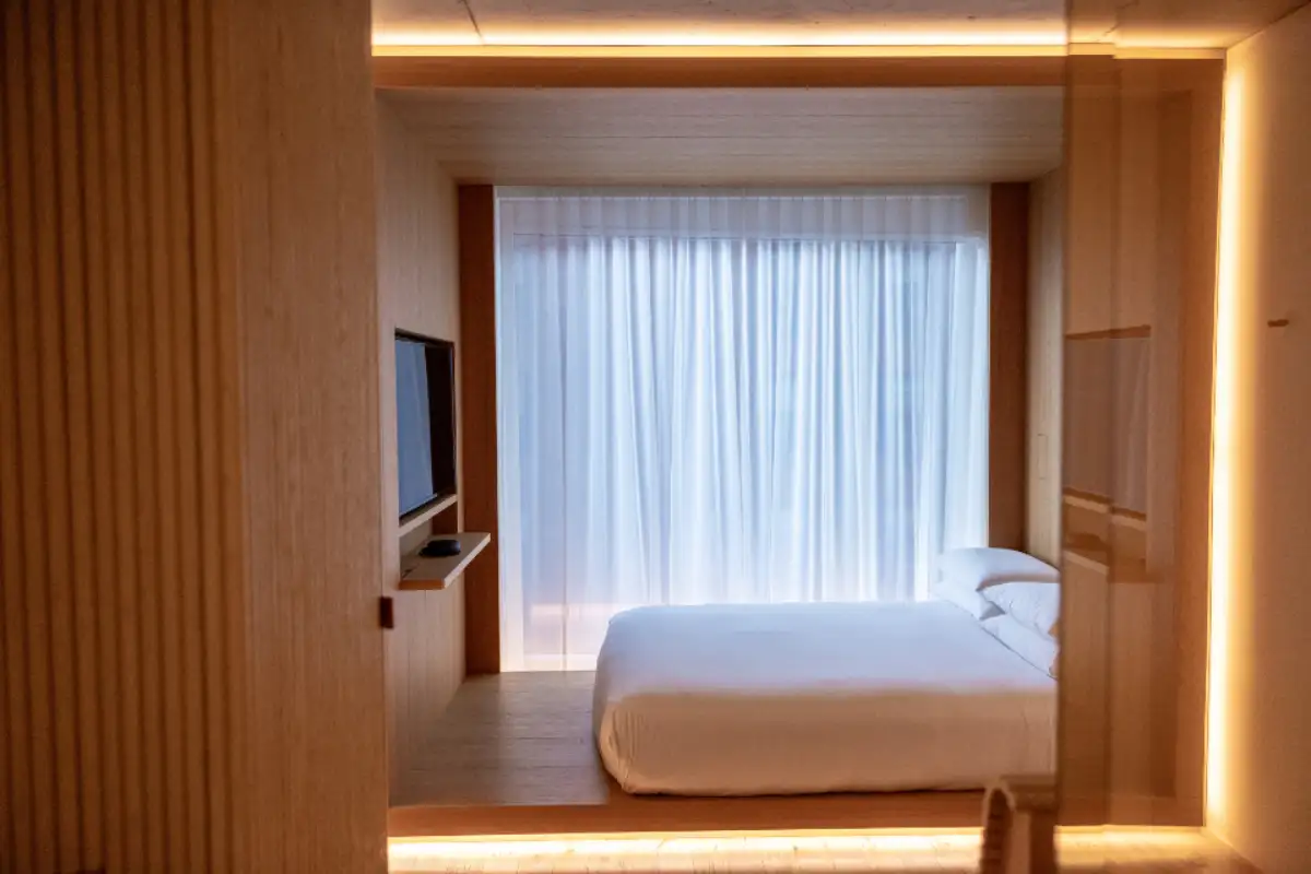 Chambre d'hôtel contemporaine habillée de bois clair, lit face à une fenêtre voilée